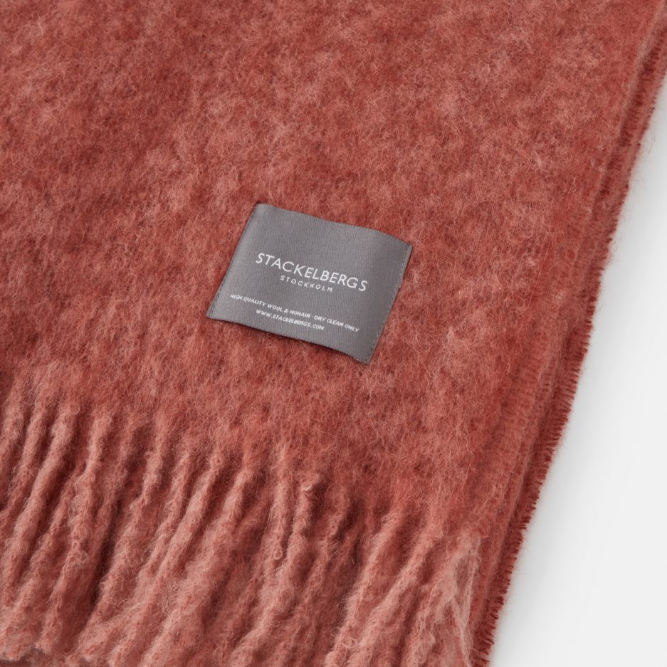 Close-up of the Stackelbergs logo on a luxurious mellow rose & brick Melange Mohair plaid on a white backdrop.