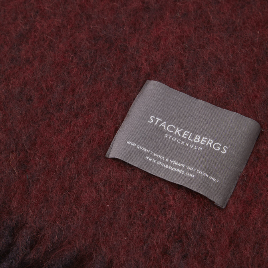 Close-up of the Stackelbergs logo on a luxurious aubergine & fired earth melange Mohair plaid on a white backdrop.