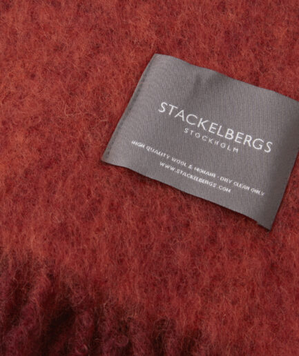 Close-up of the Stackelbergs logo on a luxurious fired earth & jaffa orange melange Mohair plaid on a white backdrop.
