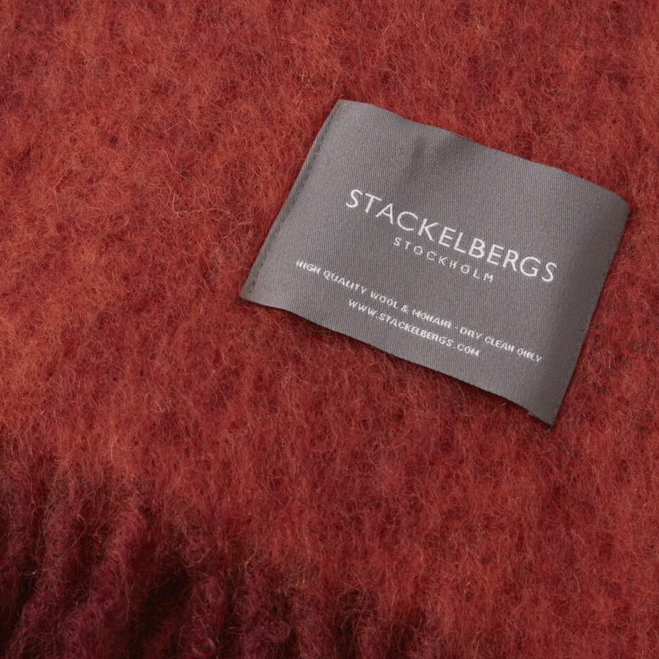 Close-up of the Stackelbergs logo on a luxurious fired earth & jaffa orange melange Mohair plaid on a white backdrop.