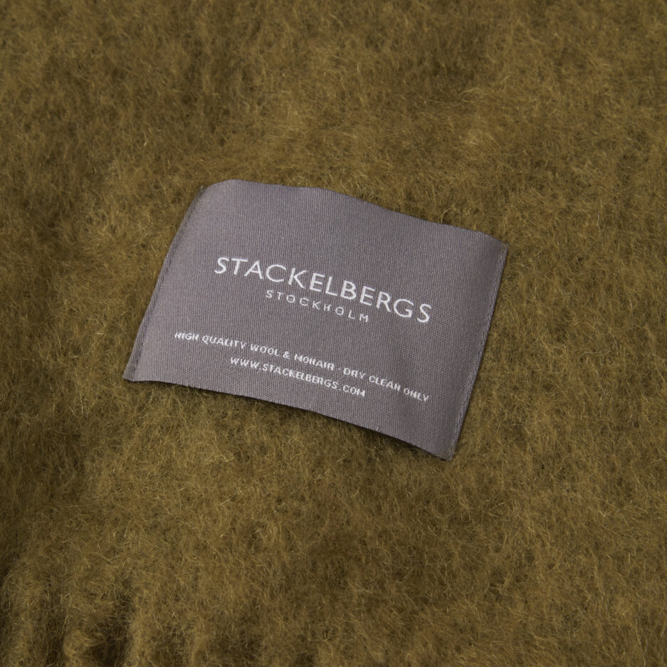 Close-up of the Stackelbergs logo on a luxurious avocado dark green Mohair plaid on a white backdrop.