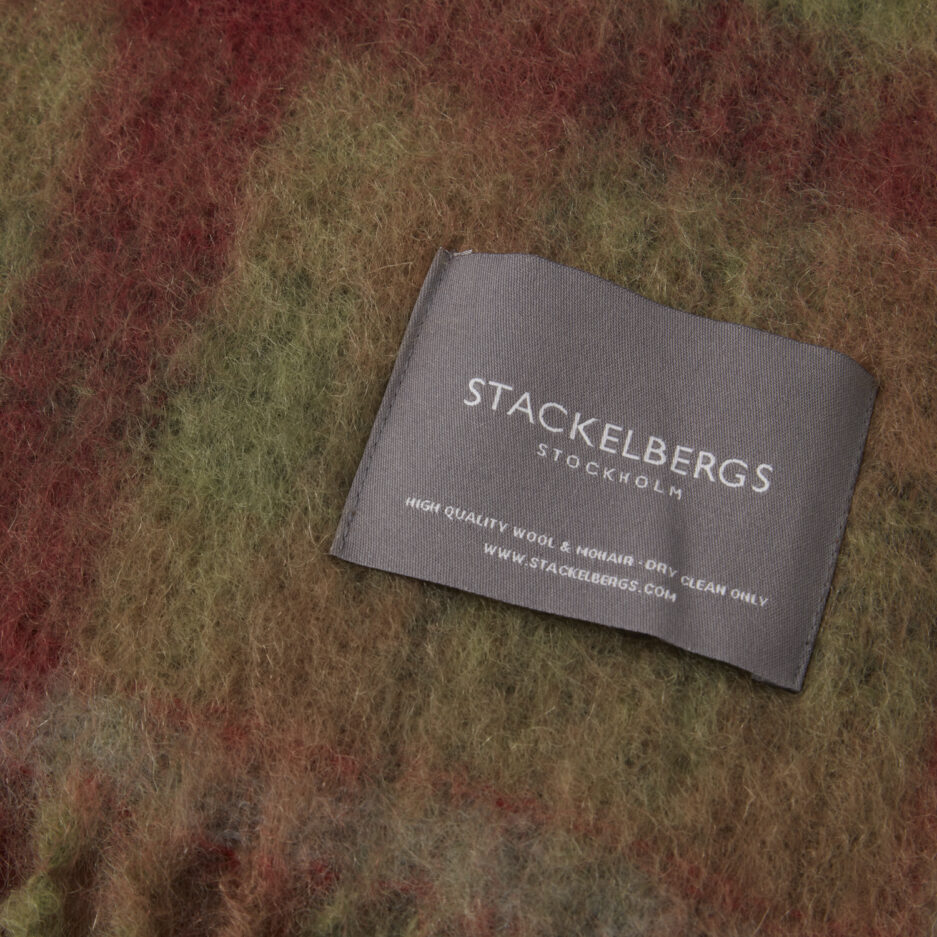 Close-up of the Stackelbergs logo on a luxurious moss & fired earth check Mohair plaid on a white backdrop.