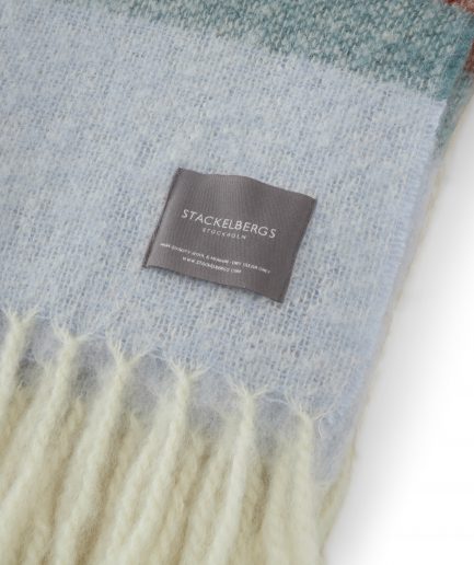 Close-up of the Stackelbergs logo on a luxurious stripe orange, mint and red Mohair plaid on a white backdrop.