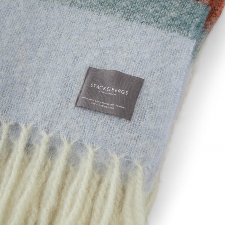 Close-up of the Stackelbergs logo on a luxurious stripe orange, mint and red Mohair plaid on a white backdrop.