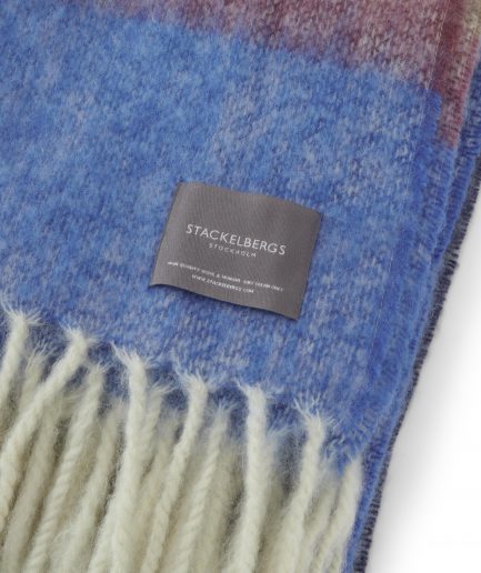 Close-up of the Stackelbergs logo on a luxurious stripe blue,azur and camel Mohair plaid on a white backdrop.