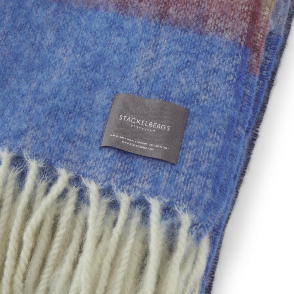 Close-up of the Stackelbergs logo on a luxurious stripe blue,azur and camel Mohair plaid on a white backdrop.