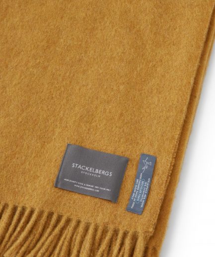 Close-up of the Stackelbergs logo on a luxurious alpaca golden yellow recycled Mohair plaid on a white backdrop.