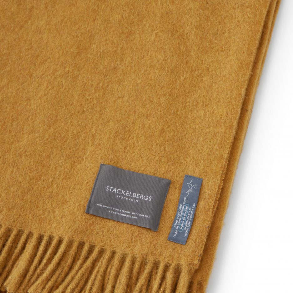 Close-up of the Stackelbergs logo on a luxurious alpaca golden yellow recycled Mohair plaid on a white backdrop.