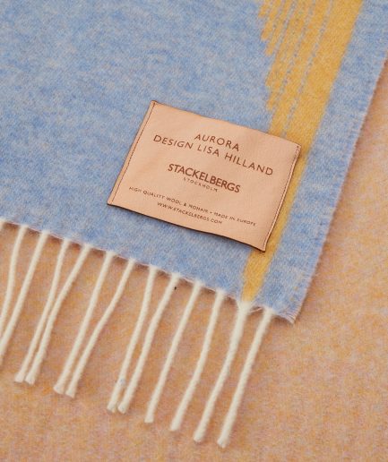 Close-up of the Stackelbergs logo on a luxurious Sky Blue, Golden Yellow & Rose wool plaid Designed by Lisa Hilland on a white backdrop.