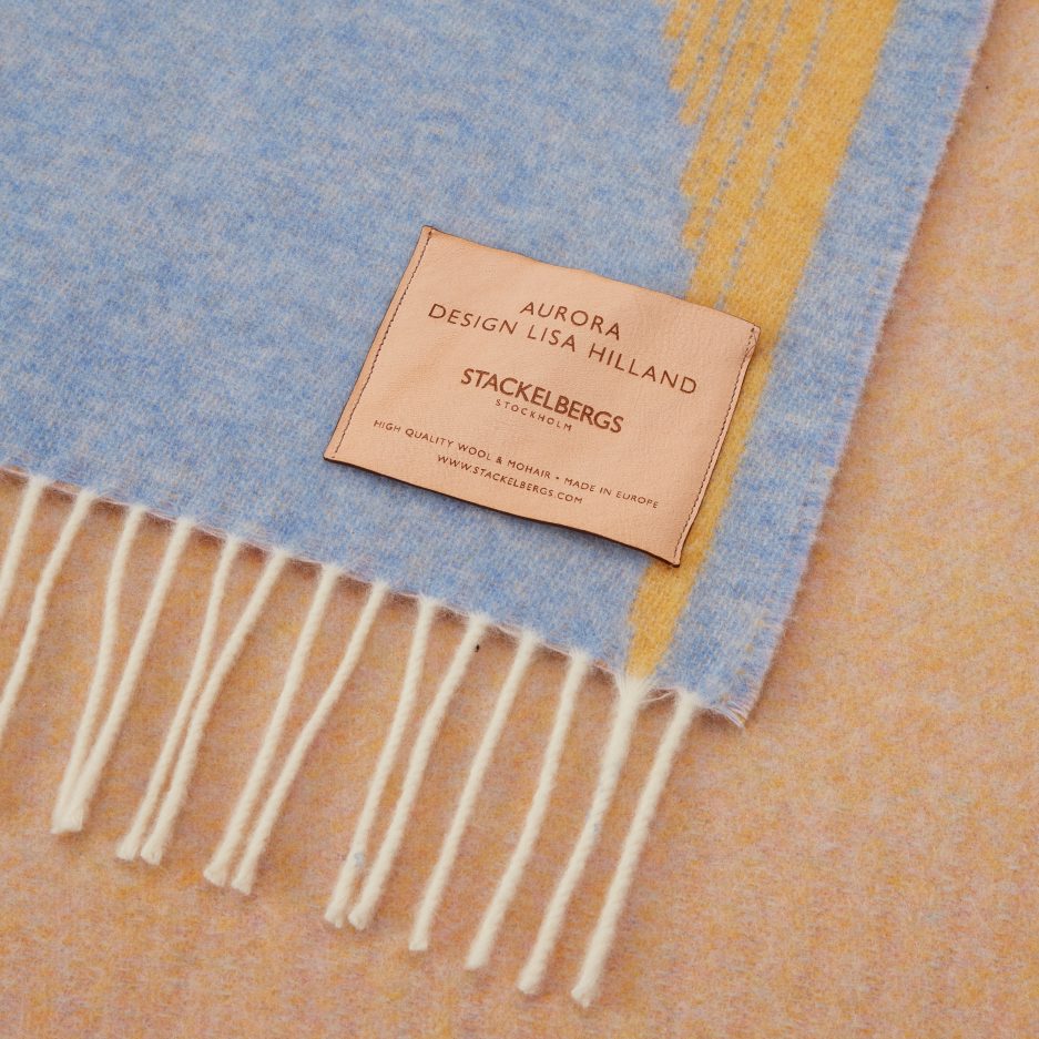 Close-up of the Stackelbergs logo on a luxurious Sky Blue, Golden Yellow & Rose wool plaid Designed by Lisa Hilland on a white backdrop.