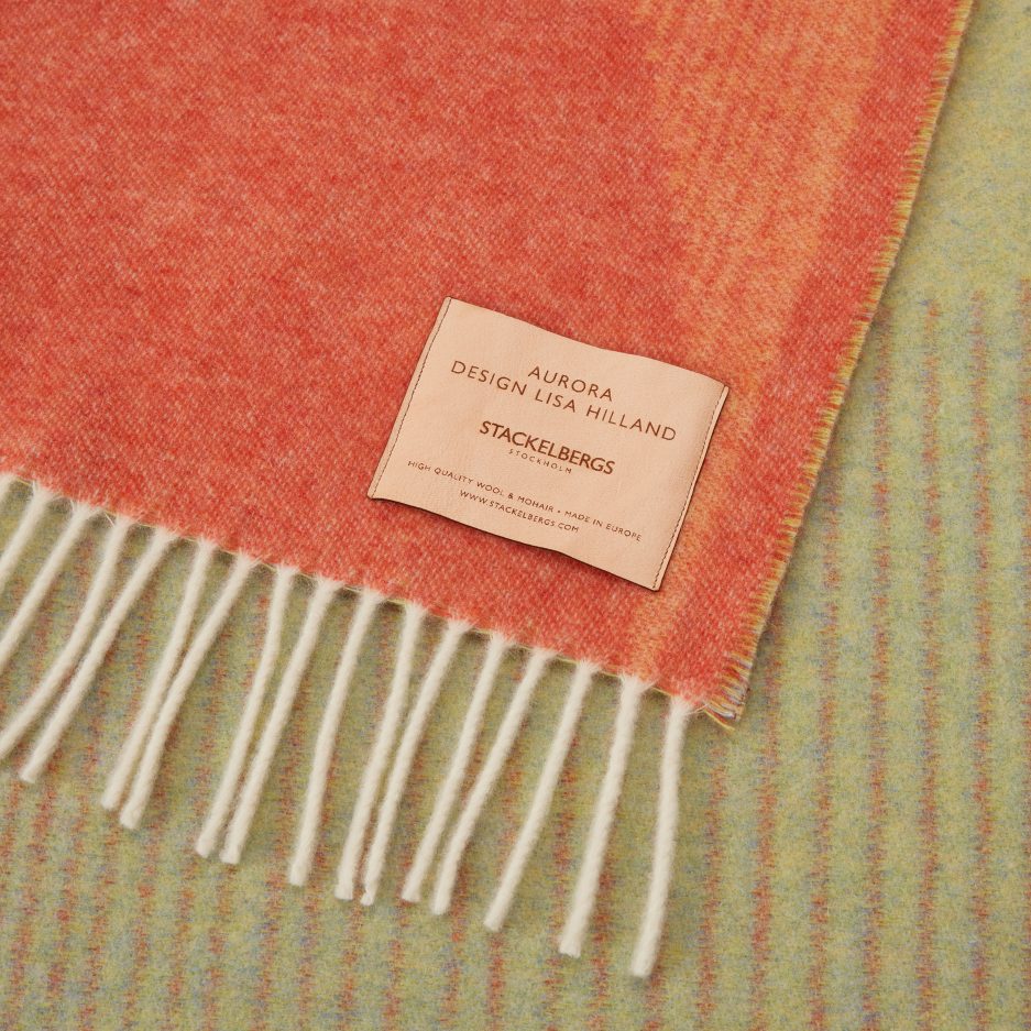 Close-up of the Stackelbergs logo on a luxurious Orange, Grass Green & Sky Blue wool plaid Designed by Lisa Hilland on a white backdrop.