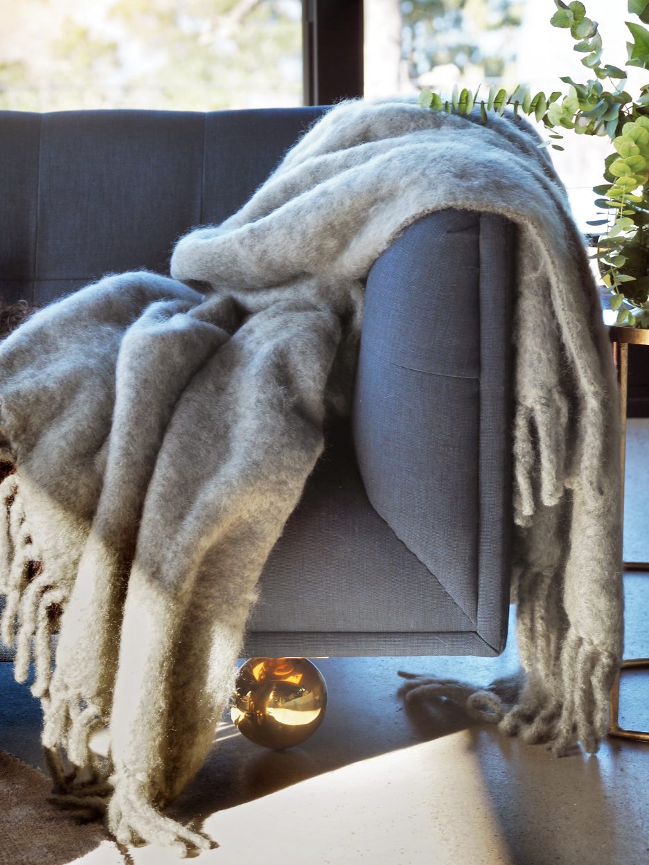 A luxurious green marble Mohair plaid partially covered in the sun hanging of a dark grey couch next to plants and a golden sphere lies underneath the couch.