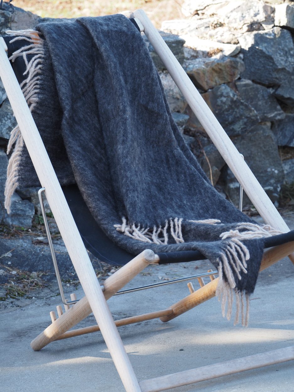 A grey luxurious Mohair plaid with beige strings laying on a wooden sun lounger with black rocks in the background.