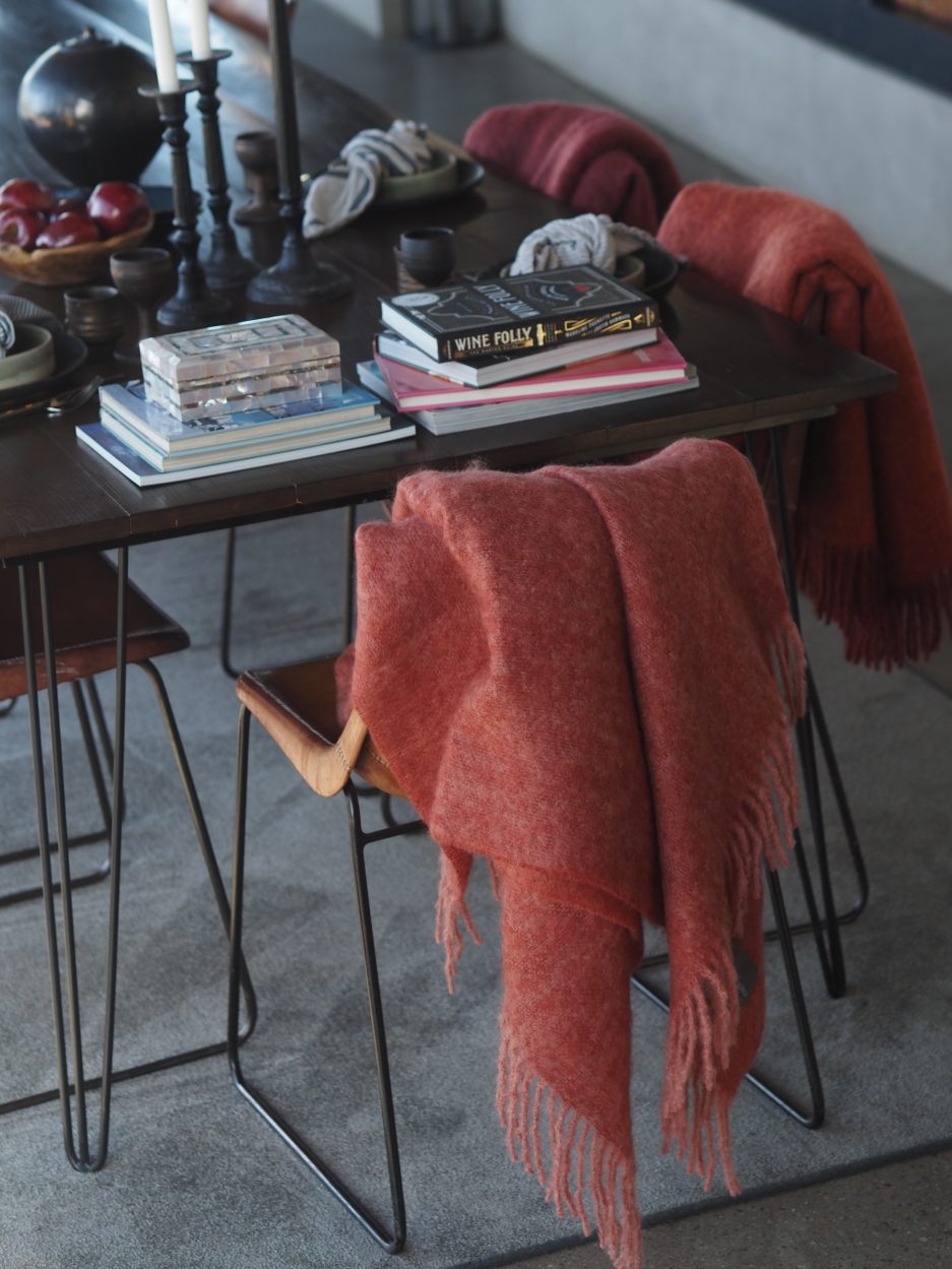 A light red luxurious Mohair plaid hangs of a modern wooden chair next to a table full of books.