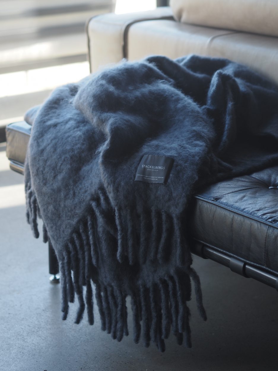 A luxurious deep blue Mohair plaid hangs of a black leather couch partially covered in the sun.